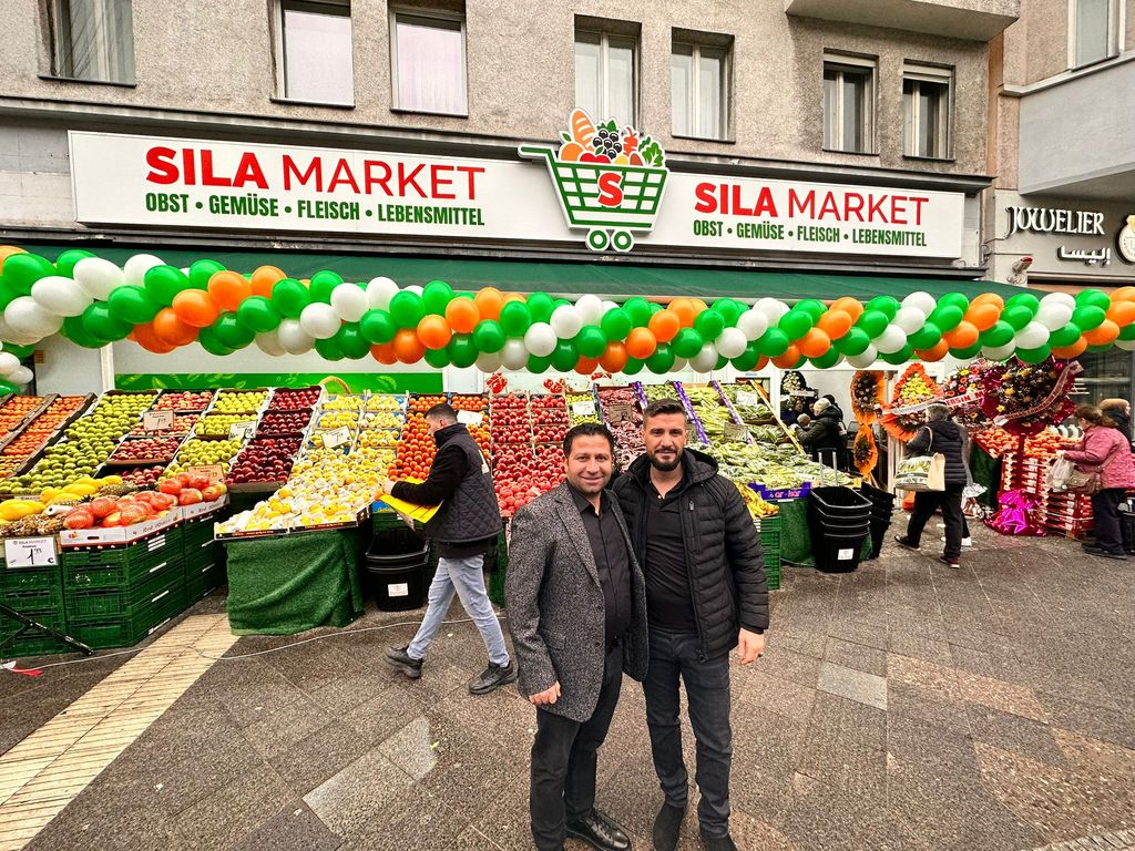 Die Ladenfront von SILA Market in der Turmstraße: Obst- und Gemüsestand unter dem grünen Vordach, davor zwei Männer aus dem Team.