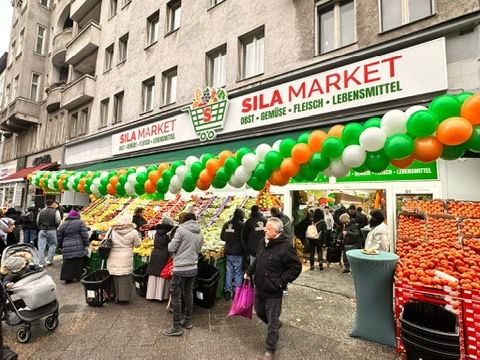 Belebter Gehweg vor SILA Market: Kundinnen und Kunden an den Obst- und Gemüsekisten, darüber das Ladenschild und grün-orange Ballons.